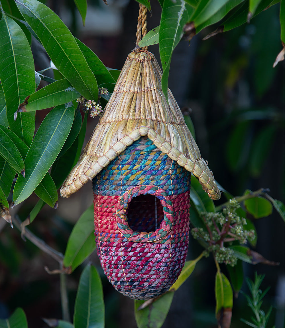 Woven birdhouse with a thatched roof hanging among green leaves